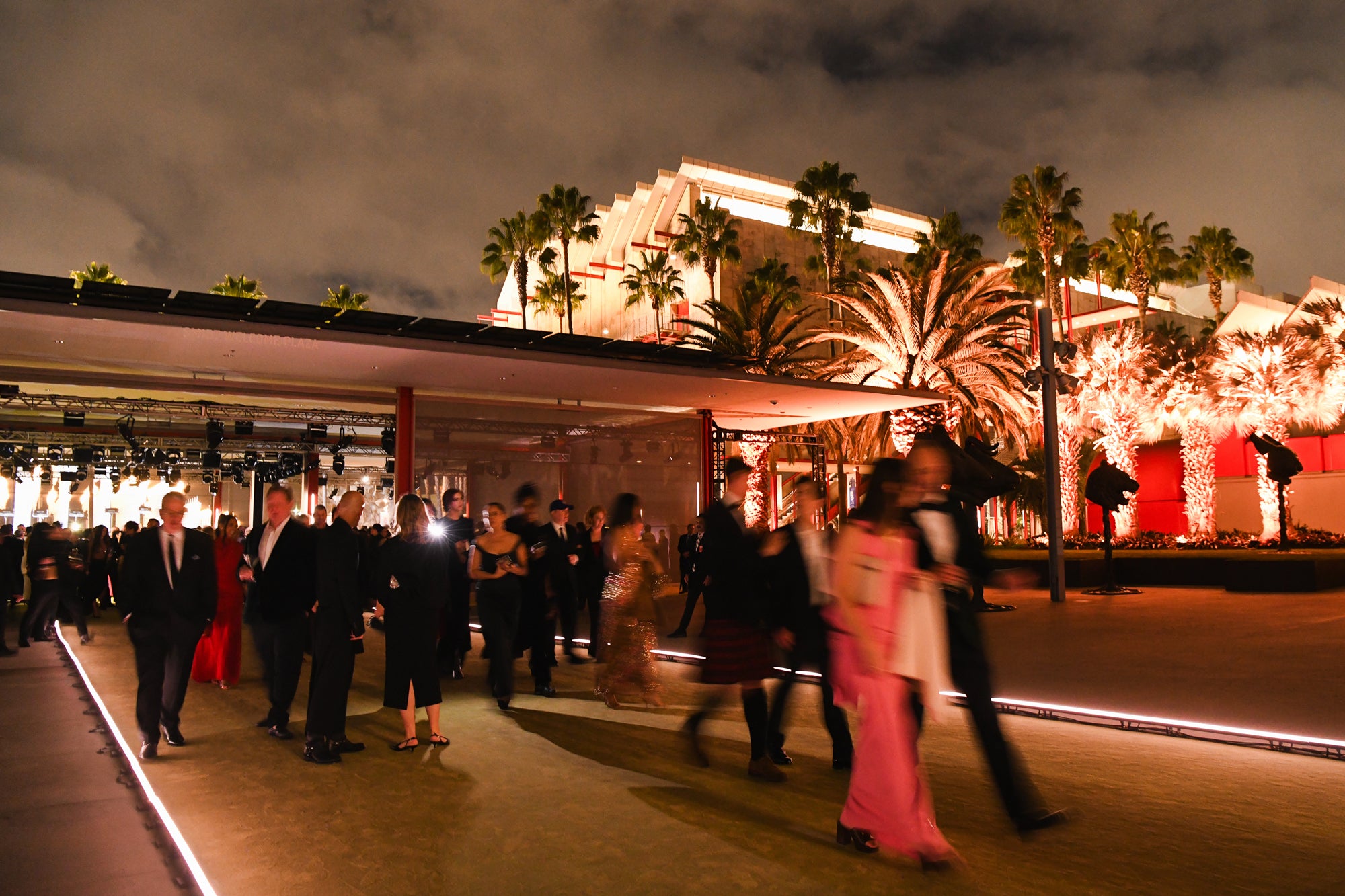 A crowd of formally dressed people walk and mingle at an evening event outside a brightly lit modern building surrounded by palm trees at LACMA. The scene is illuminated with warm lighting, and the sky above is overcast.