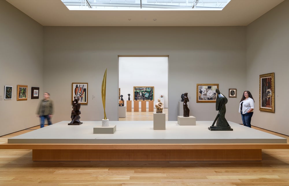 A museum gallery with high ceilings and skylights displays a central platform of modernist sculptures, surrounded by framed paintings on neutral-colored walls. 