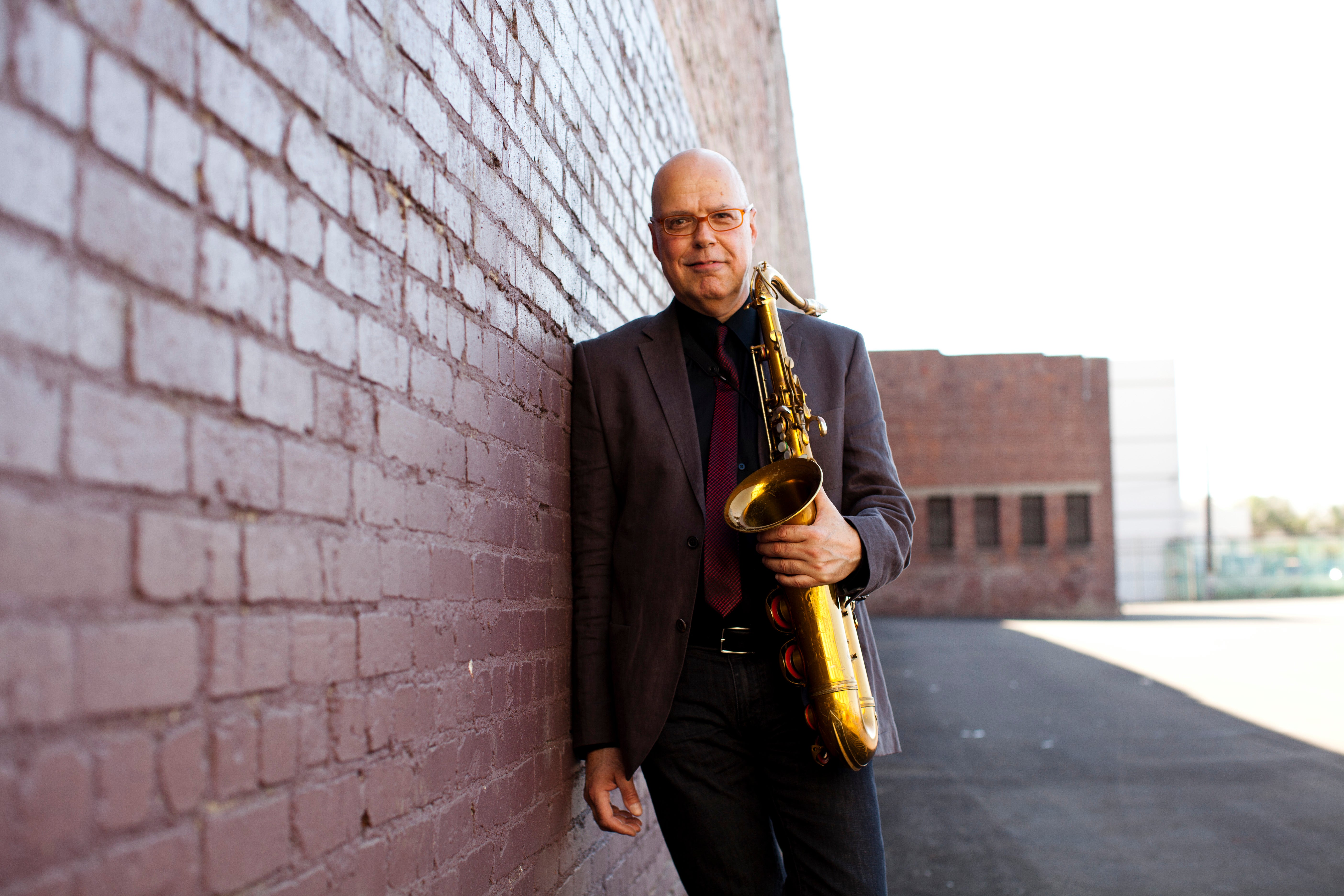 Bob Sheppard leaning against a brick wall holding a saxophone