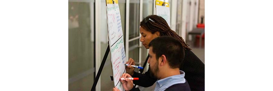 Two people sitting in front of a whiteboard, one is writing at the top of the whiteboard, the other is writing on the bottom of the whiteboard