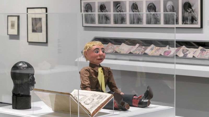 A ventriloquist dummy in a brown shirt, yellow tie, and cowboy boots sits next to an open book and a black leather bust in a vitrine