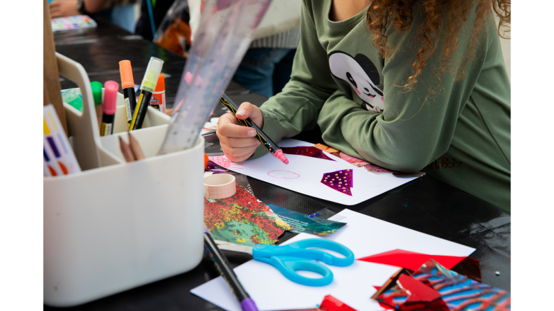child drawing on paper using a paint marker, surrounded by art materials including decorative paper, scissors, paint markers, and tape
