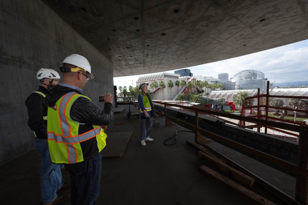 A man in safety gear viewing the vista from an opening of a gray building under construction, with two people observing him in the foreground